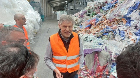 A group of people wearing orange safety vests stands next to large piles of sorted plastic waste inside a recycling facility. One man is smiling and speaking to others in the group.
