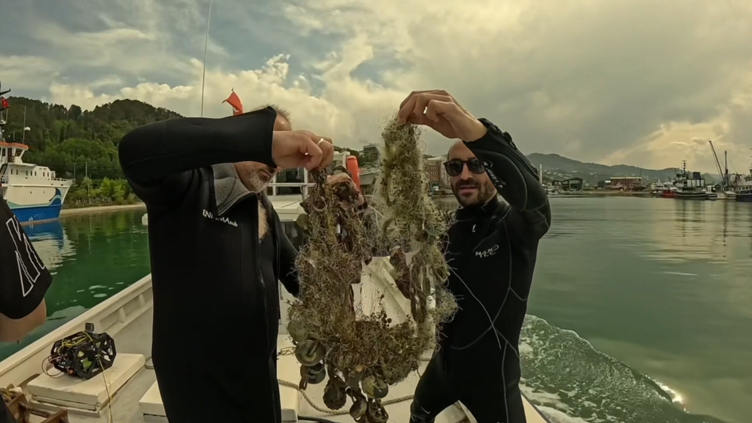 Two people in wetsuits on a boat hold up a tangled mass of seaweed and debris, possibly collected from the water. Boats and a shoreline with trees and mountains are visible in the background under a partly cloudy sky.