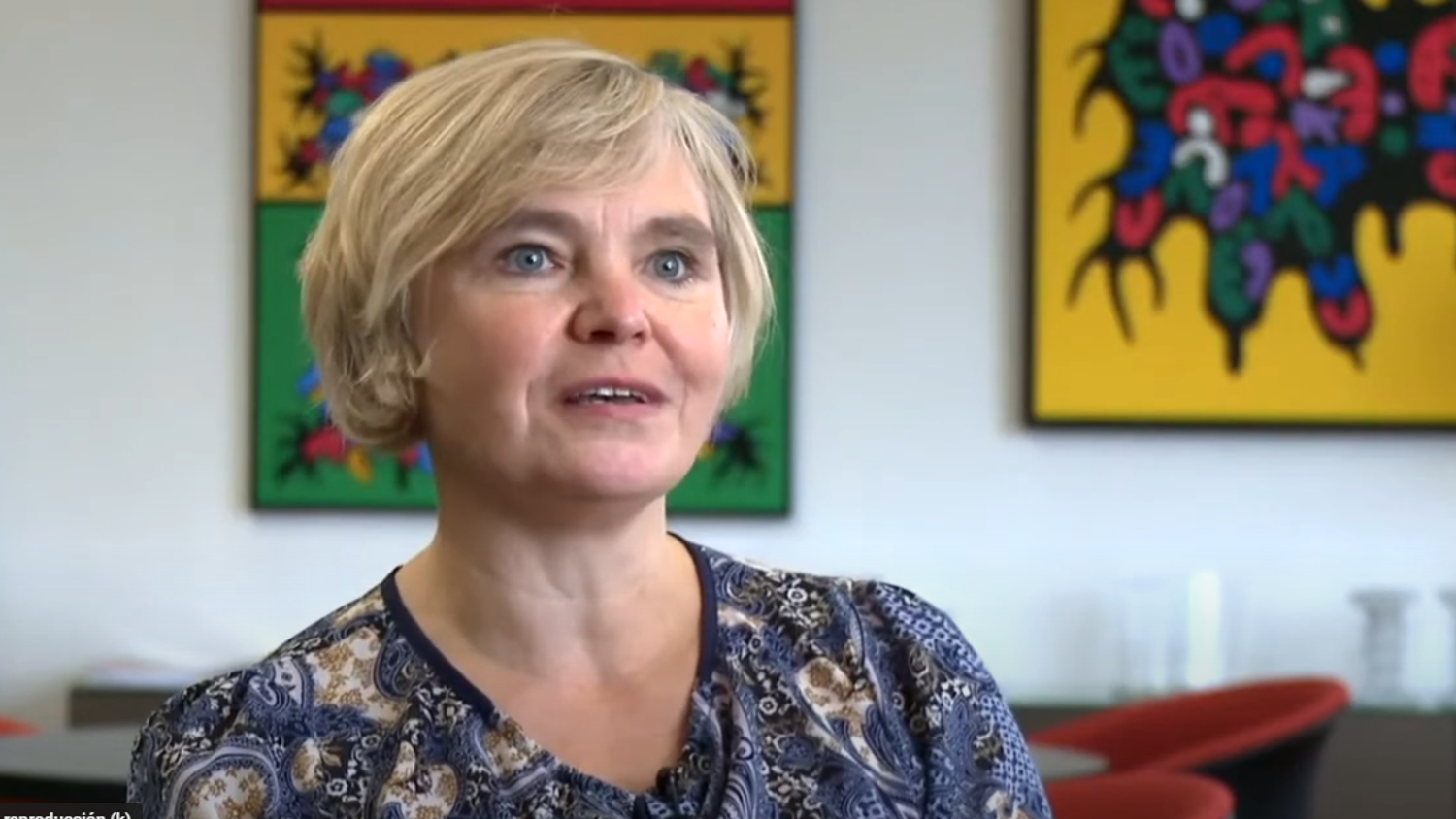 A woman with short blonde hair wearing a patterned blouse is sitting in a room with colorful abstract art on the walls.