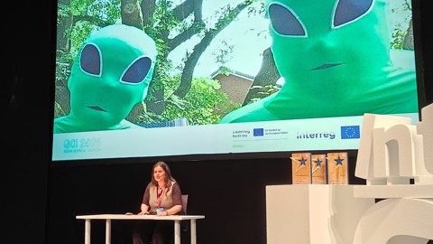 A woman sits at a desk on stage, speaking, with a large screen behind her showing two people in green alien costumes with big black eyes standing outdoors.