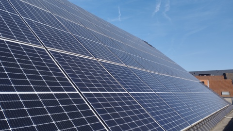 A close-up view of large solar panels installed on a rooftop with a clear blue sky and rooftops of nearby buildings in the background.