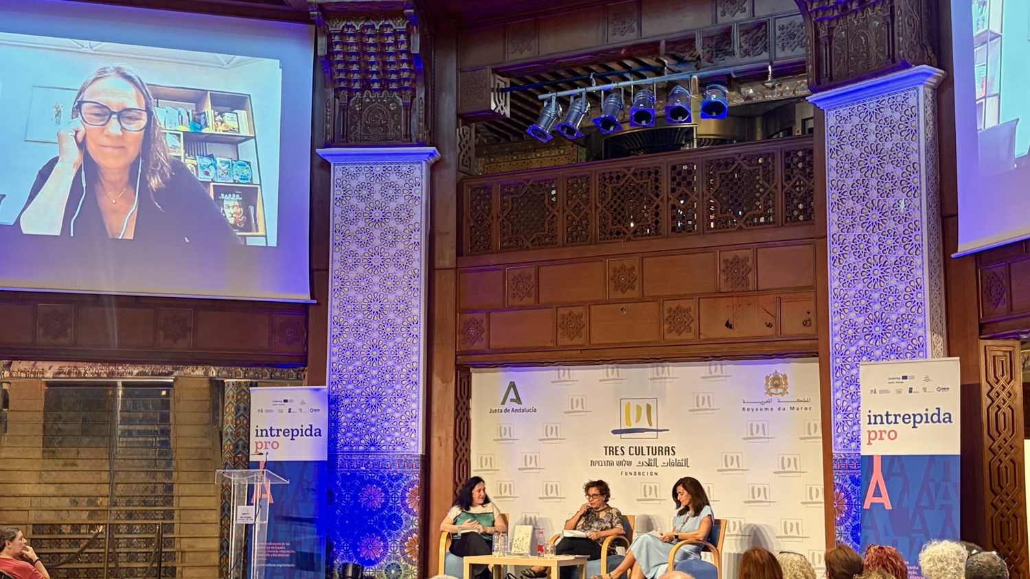 Three women sit on a stage during a panel discussion in a wood-paneled room, with a large screen behind them showing a remote speaker.