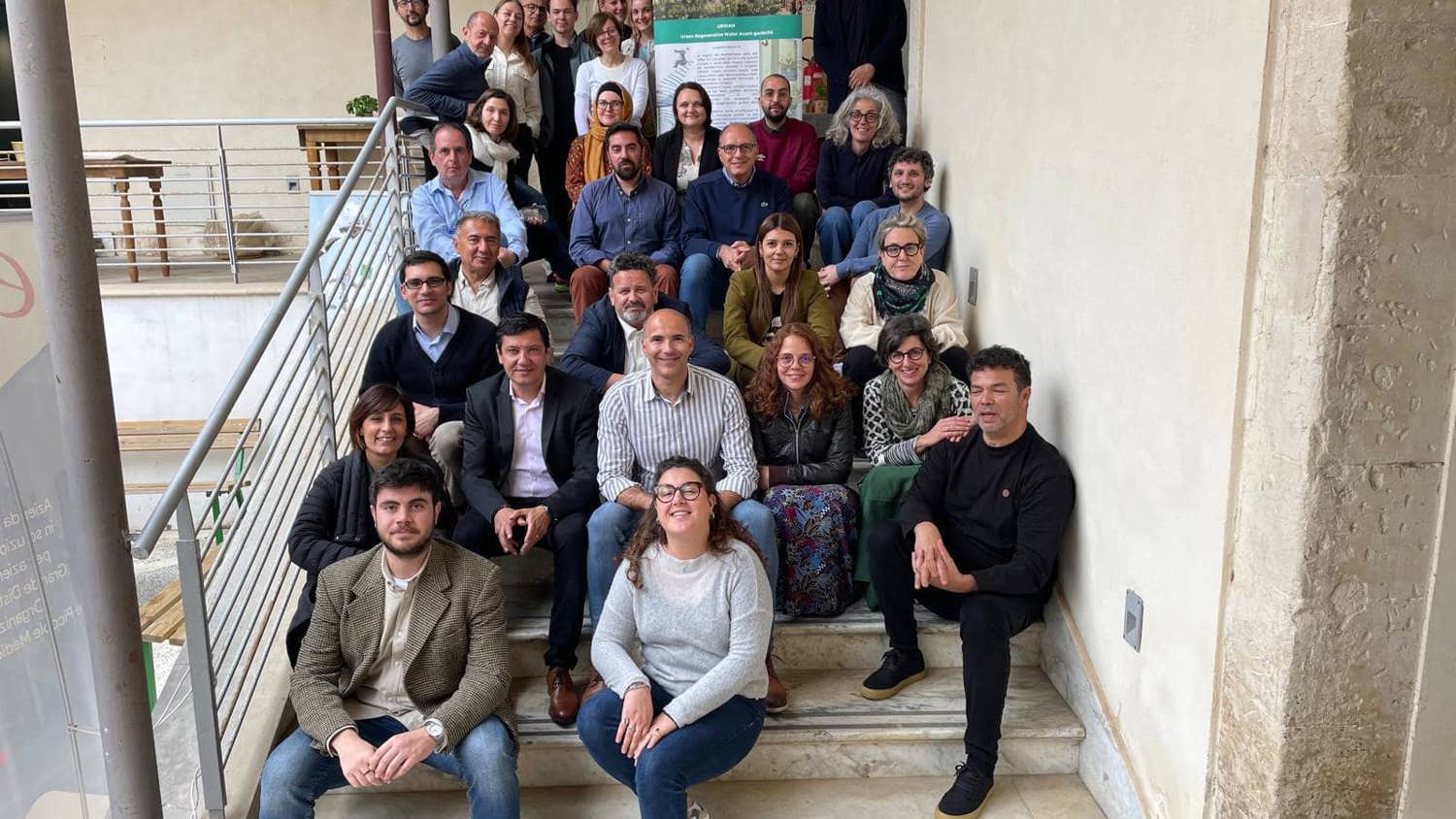 A group of people posing on stairs in an indoor setting with a poster in the background. They appear to be at an event or gathering, wearing casual to business casual attire. The setting includes a railing and beige walls.