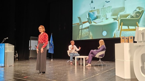 A woman stands on stage while two people sit and talk near a small table. A large screen behind them displays a modern living room scene. The setting appears to be a formal presentation or discussion event.