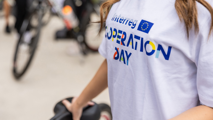 Person wearing a white shirt with "Interreg Cooperation Day" printed on it, holding a bicycle saddle. Blurred bicycles and people are in the background.