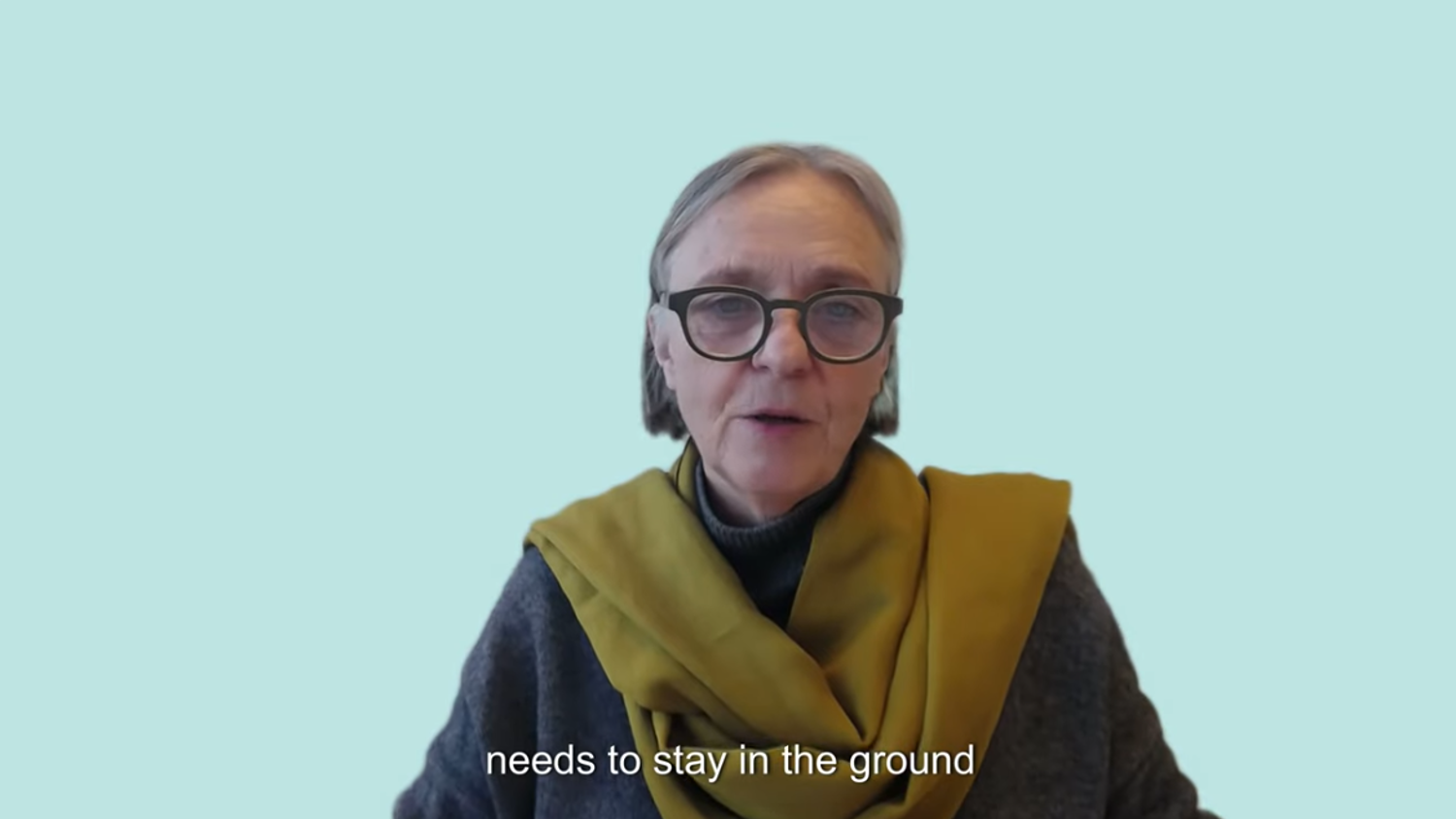 An older woman with glasses and short gray hair speaks against a light blue background, wearing a gray sweater and a mustard yellow scarf. Subtitle reads: "needs to stay in the ground".