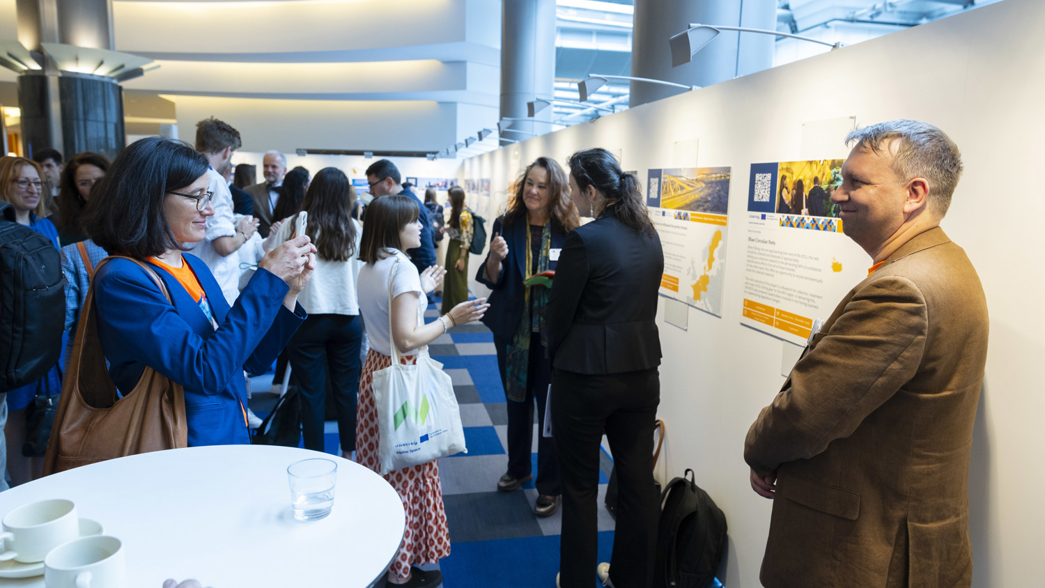 A group of people interact and view posters in a brightly lit exhibition space. A woman takes a photo with her phone while others converse and look at displays on the walls.