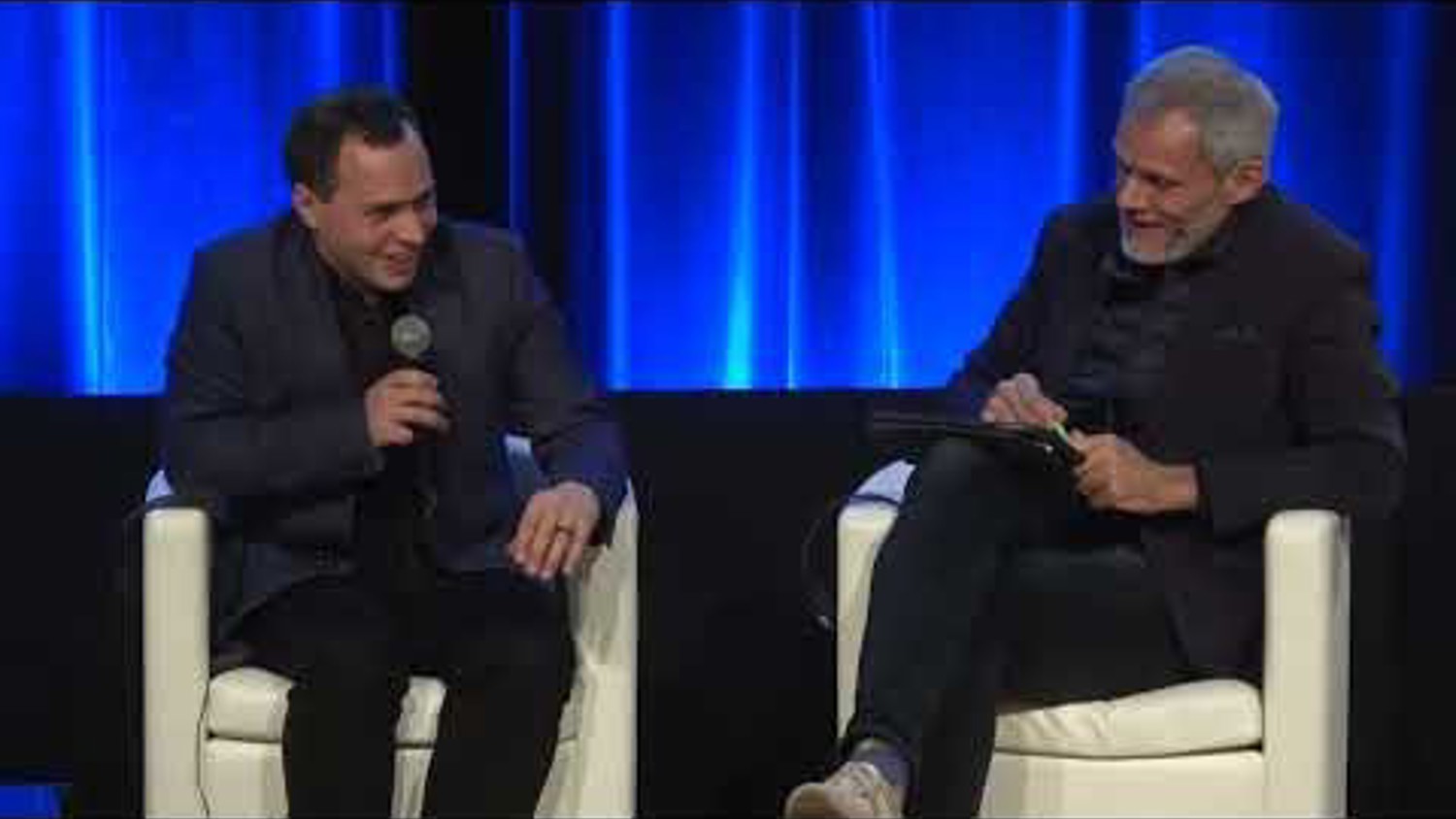 Two men sitting in white chairs on a stage with a blue backdrop. The man on the left holds a microphone and smiles. The man on the right writes on a notepad.