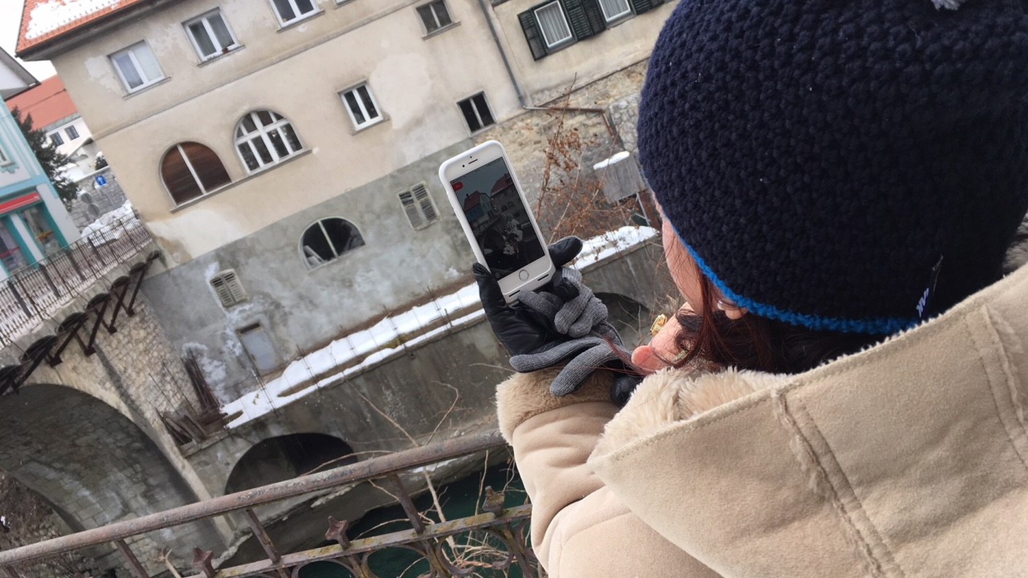 A person in a winter coat and beanie takes a selfie with a smartphone while standing on a bridge. In the background, snow-dusted buildings line the riverbank, and part of an arched bridge is visible.