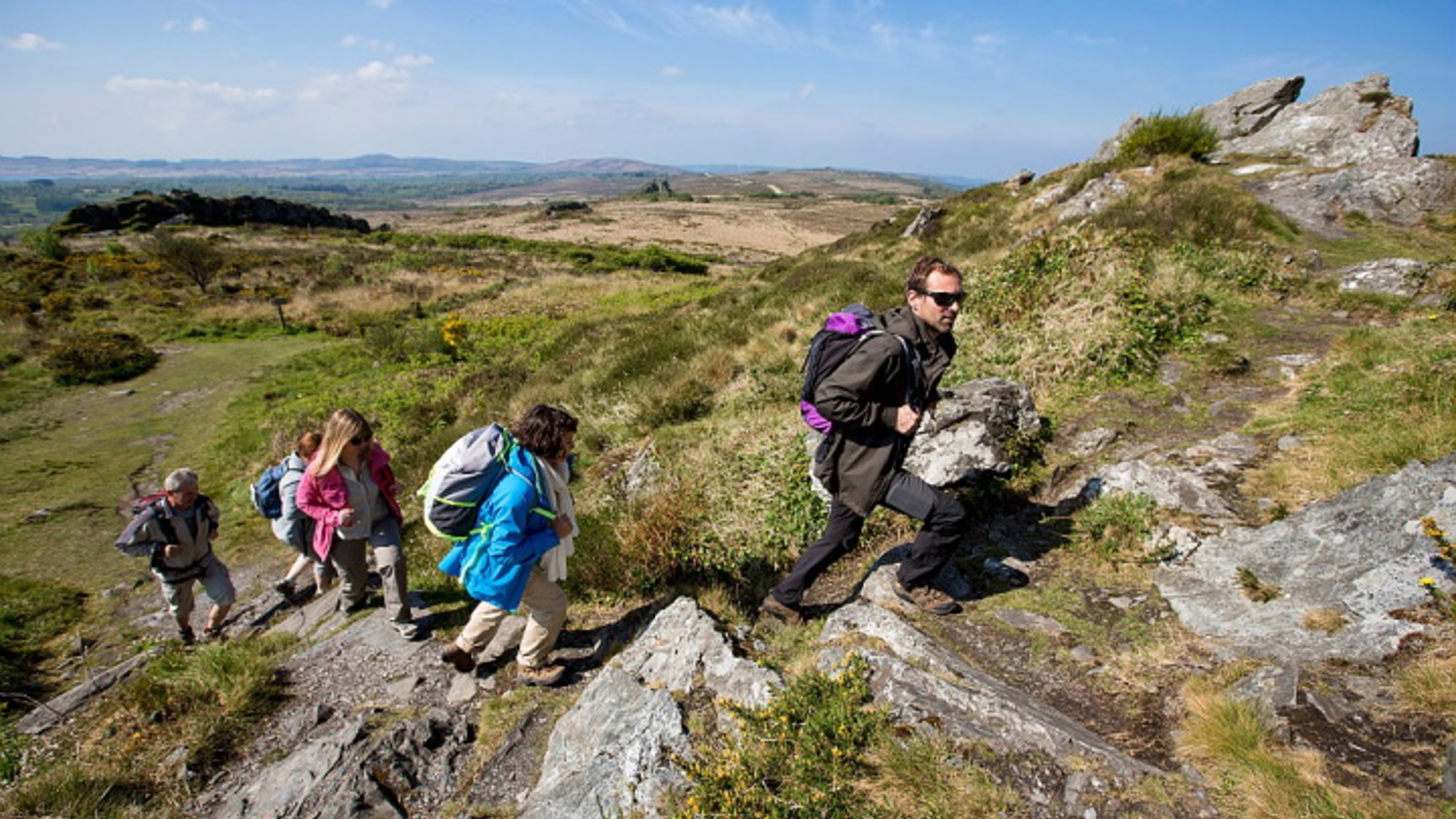 A group of hikers with backpacks traverse a rocky trail on a grassy hillside, under a clear blue sky. The landscape in the background features rolling hills and sparse vegetation.