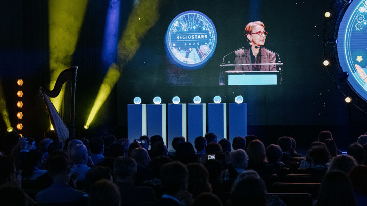 A woman speaks at a podium onstage during an event, with a "REGIOSTARS 2023" logo displayed on a large screen behind her. The audience faces the stage, and five trophy-like objects are lined up beside her.