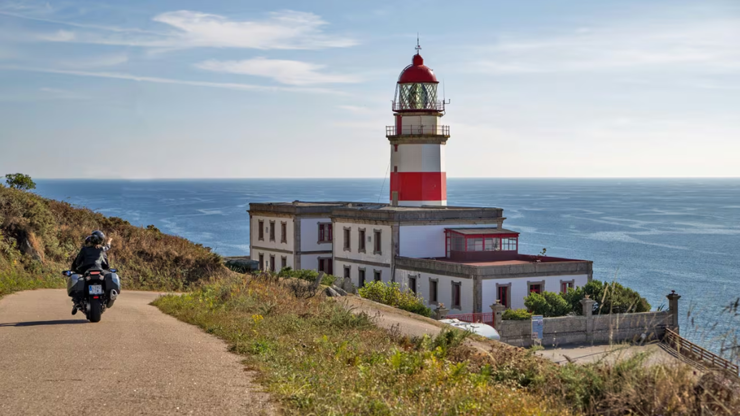 A person rides a motorcycle down a coastal road toward a white and red lighthouse overlooking the blue sea under a clear sky.