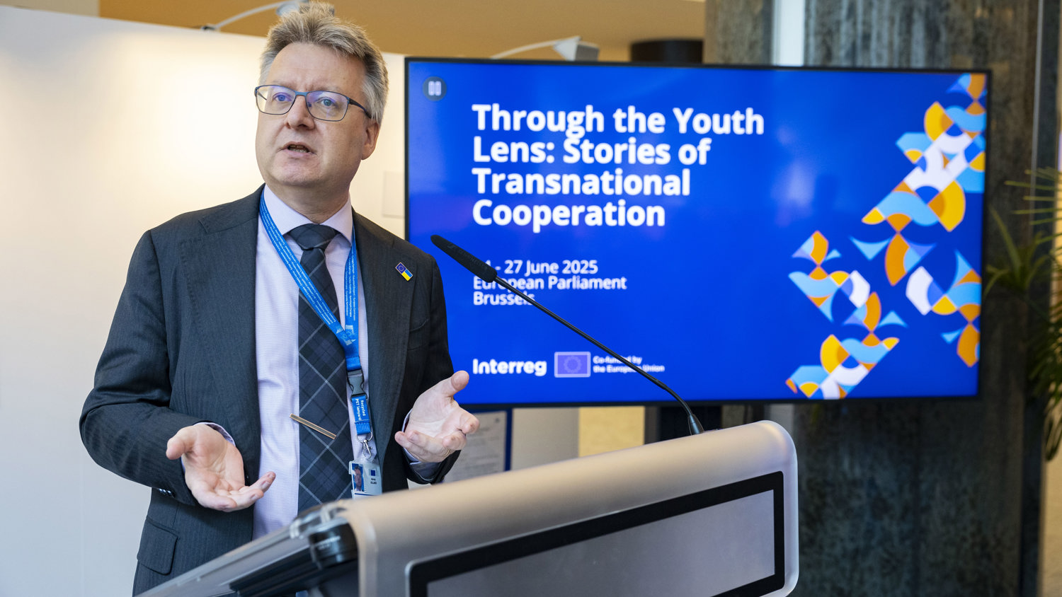 A man in a suit speaks at a podium beside a screen displaying the text "Through the Youth Lens: Stories of Transnational Cooperation, 26-27 June 2025, European Parliament, Brussels.