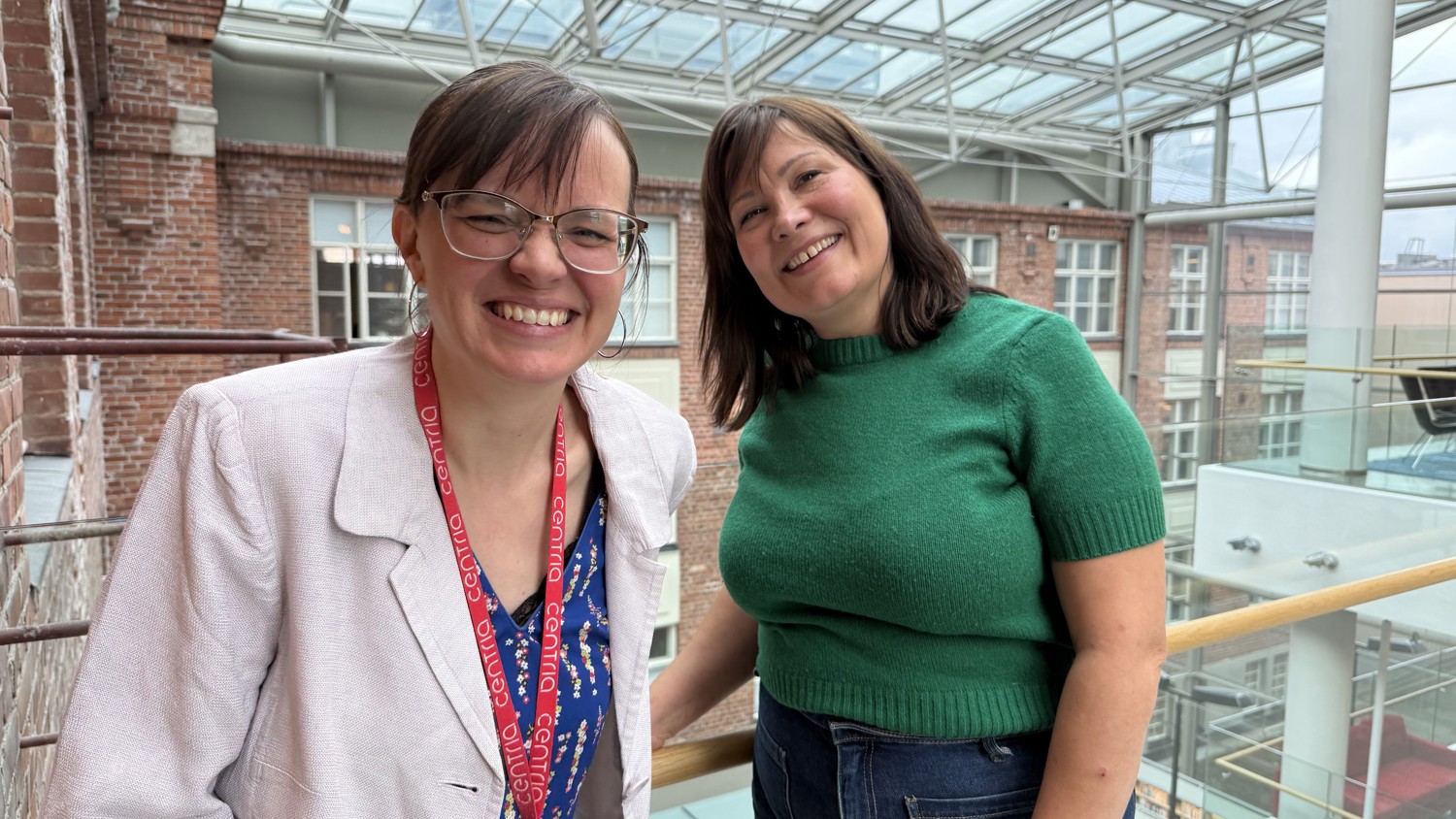 Two women smiling indoors near a glass railing, with brick walls and large windows in the background under a glass ceiling.