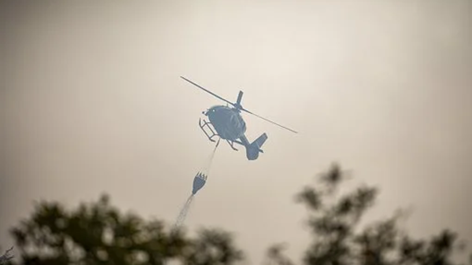 Helicopter using a water bucket to extinguish a fire, flying over treetops with a hazy sky in the background.
