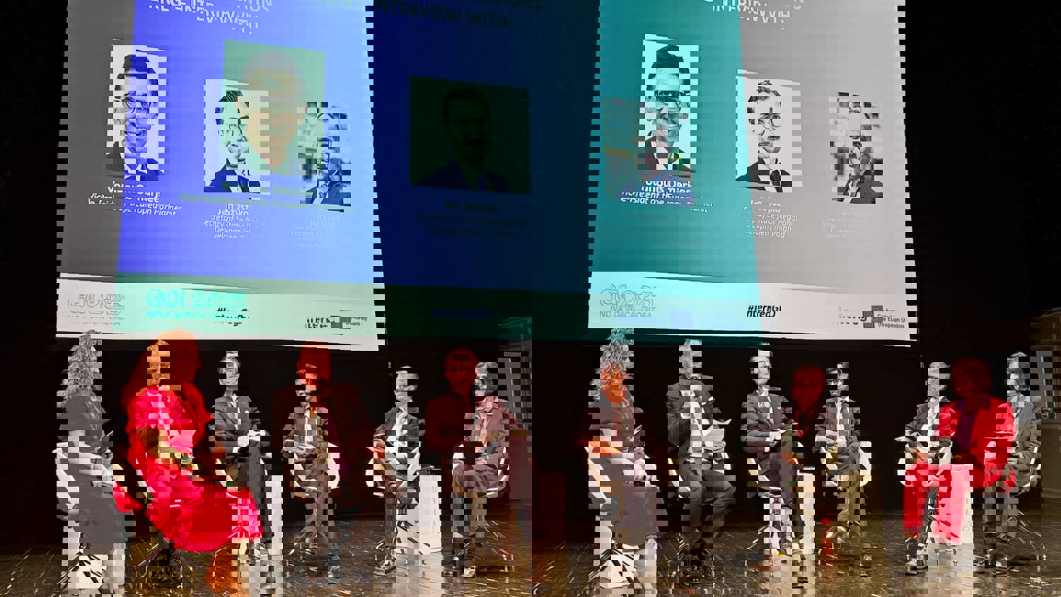 A group of people sits around a conference table in a modern office, engaged in a meeting. Large windows in the background let in natural light, and there are laptops and papers on the table.