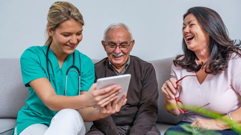 A female healthcare worker in scrubs shows a tablet to a smiling elderly man and a laughing woman sitting on a couch. The group appears to be having an enjoyable and engaging conversation.