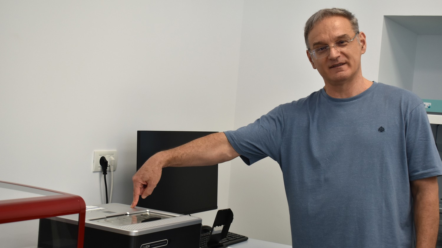 A man wearing glasses and a blue t-shirt points to a laboratory instrument on a desk.