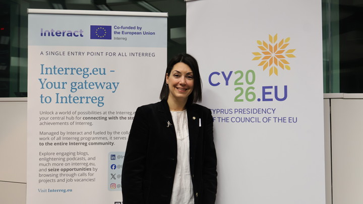 A woman in a black suit stands smiling between two banners: one for Interreg.eu and one for CY2026.EU, representing Cyprus’s presidency of the Council of the EU. The setting appears to be an indoor event or conference.