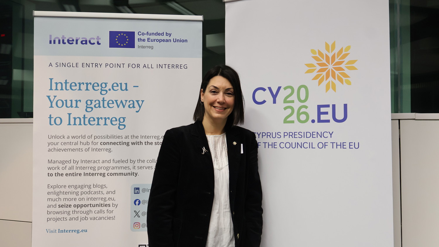 A woman in a black suit stands smiling between two banners: one for Interreg.eu and one for CY2026.EU, representing Cyprus’s presidency of the Council of the EU. The setting appears to be an indoor event or conference.