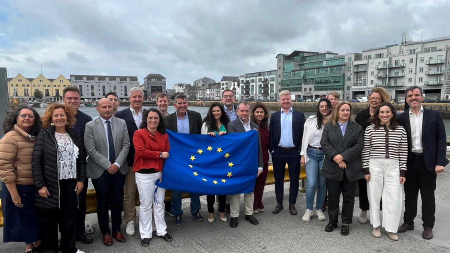 A group of about 20 people stand outdoors by a waterfront, holding a European Union flag. Modern buildings and cloudy skies are in the background. The group is dressed in business casual attire and smiling at the camera.