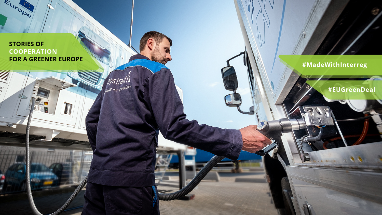 A worker operates a fueling station, handling a hose connected to a vehicle. The backdrop shows trailers with "Stories of Cooperation for a Greener Europe" and hashtags #MadeWithInterreg and #EUGreenDeal featured on the image.