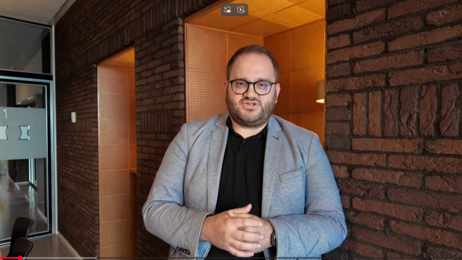 A man with glasses and a beard, wearing a gray blazer and black shirt, stands indoors against a brick wall, speaking and gesturing with his hands. The setting appears to be an office or modern hallway.
