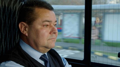 A man wearing a blue shirt and tie, with a dark vest, sits in a vehicle looking out the window. The scene outside is blurred, indicating movement. He appears thoughtful and focused.