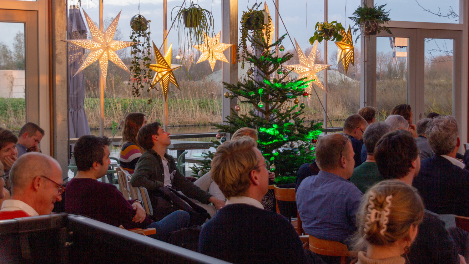 A group of people sits indoors, facing forward, in a room decorated with glowing star-shaped lights, hanging plants, and a Christmas tree with green lights near large windows. It’s daytime outside.