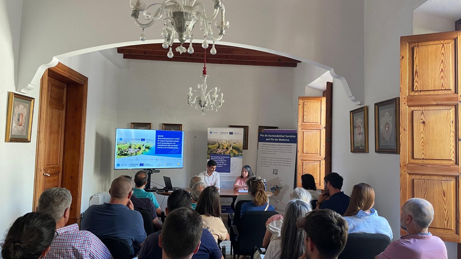 A group of people sit facing two speakers at the front of a bright room during a presentation. A TV screen displays slides, and wooden doors and windows line the walls. A chandelier hangs from the ceiling.