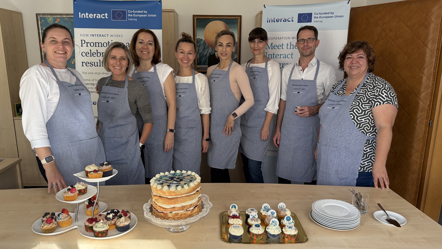 Eight people wearing aprons stand behind a table with a layered cake, cupcakes, and pastries. Interact banners are displayed in the background. The group is smiling and posing for the photo indoors.