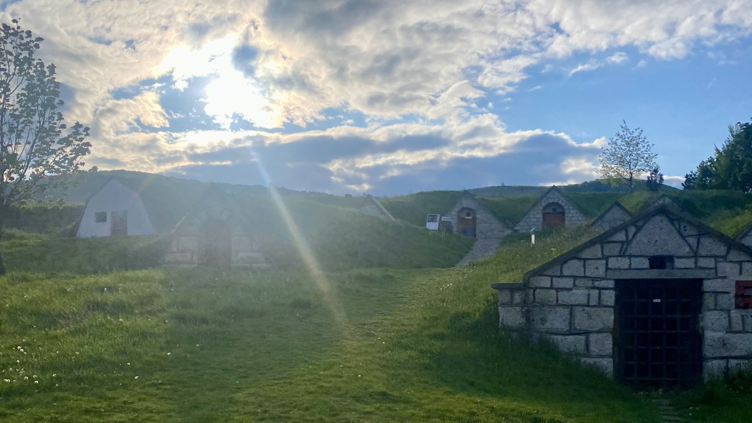 Grassy hillside with several small, stone and brick structures built into the slope, under a partly cloudy sky with the sun shining through, casting long shadows.