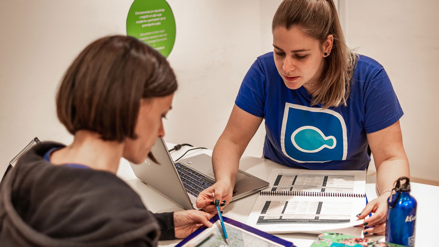 Two women sit at a table with a laptop, planner, and documents. One woman is pointing at a piece of paper while the other listens attentively. Both appear focused and are engaged in a discussion.