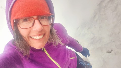 A person wearing glasses, a red beanie, purple jacket, and gloves smiles while standing in the snow. The background is snowy and white, suggesting a cold, wintry environment.