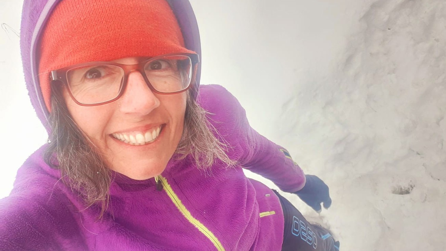 A person wearing glasses, a red beanie, purple jacket, and gloves smiles while standing in the snow. The background is snowy and white, suggesting a cold, wintry environment.