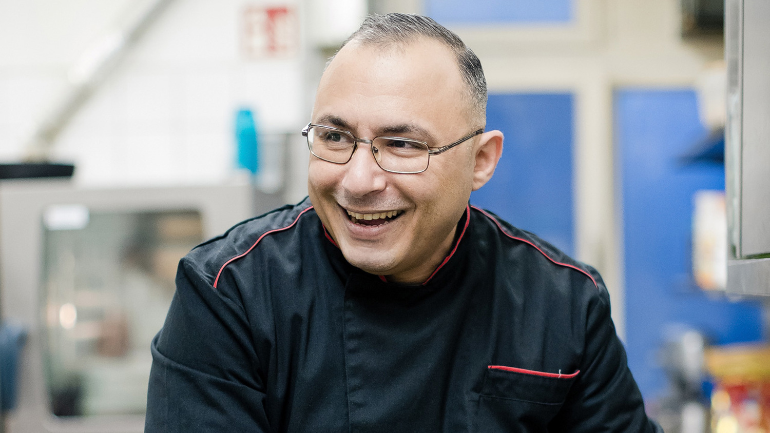 A smiling chef with glasses and a black jacket prepares food in a kitchen, slicing tomatoes on a green cutting board. He wears a red apron and white glove, surrounded by kitchen equipment and ingredients.