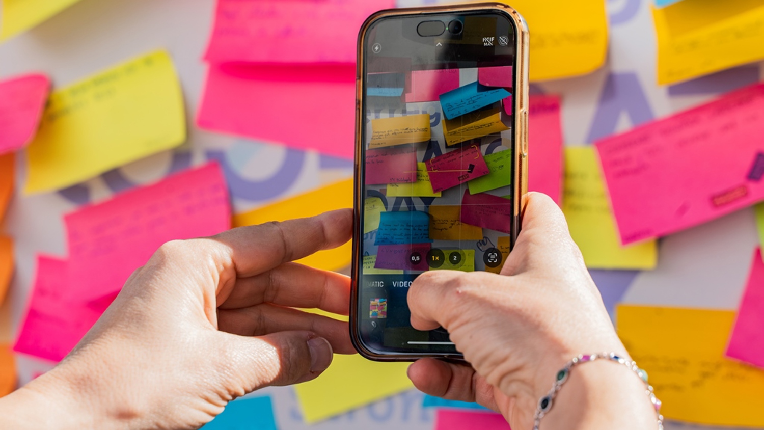 A person holds a smartphone and takes a photo of a wall covered with colorful sticky notes, some of which have handwritten messages. The phone screen displays a close-up of the notes.