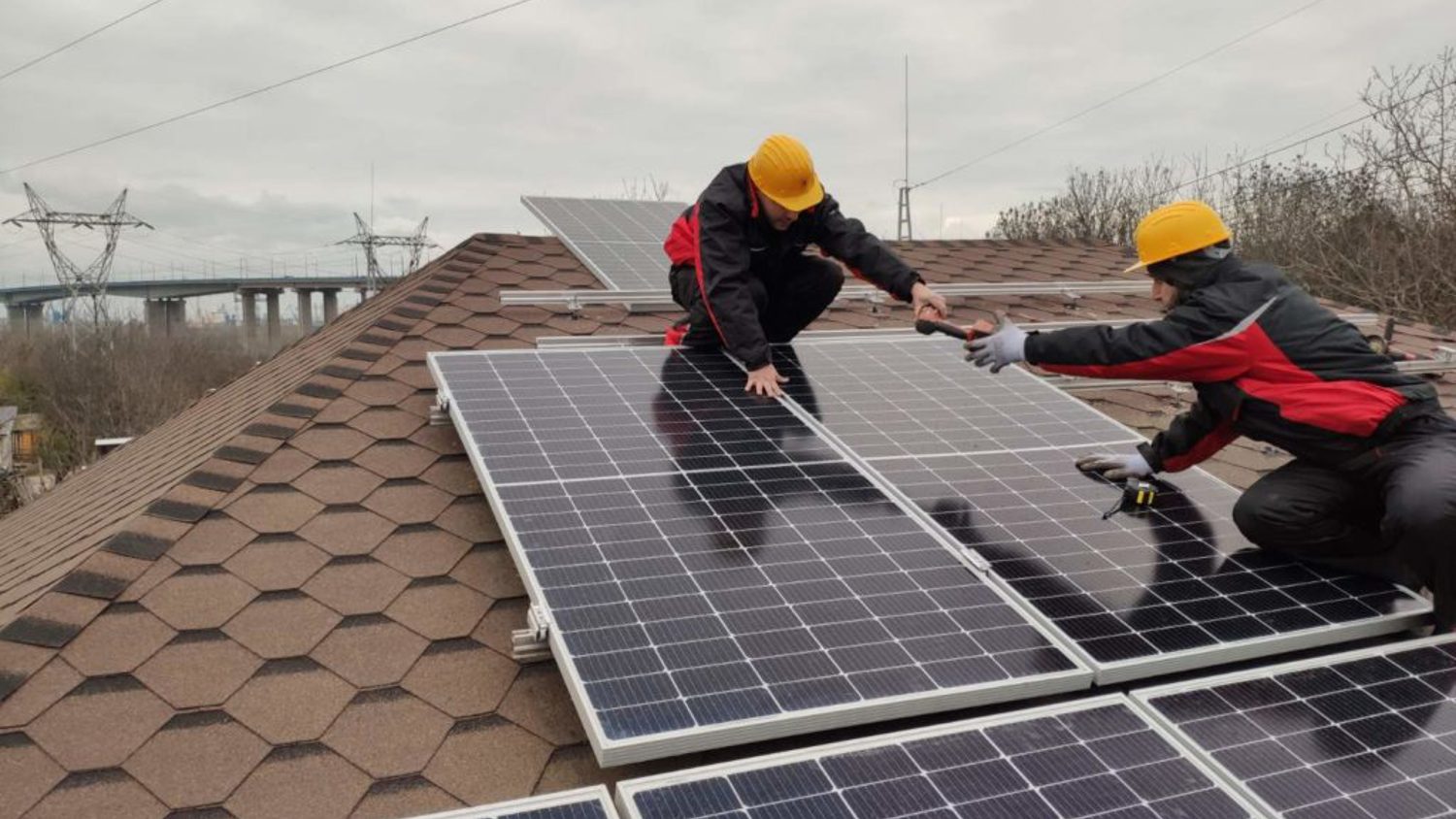 Two workers in yellow helmets and red jackets install solar panels on a sloped shingle roof. One holds a screwdriver while adjusting a panel. Power lines and a bridge are visible in the background against a cloudy sky.