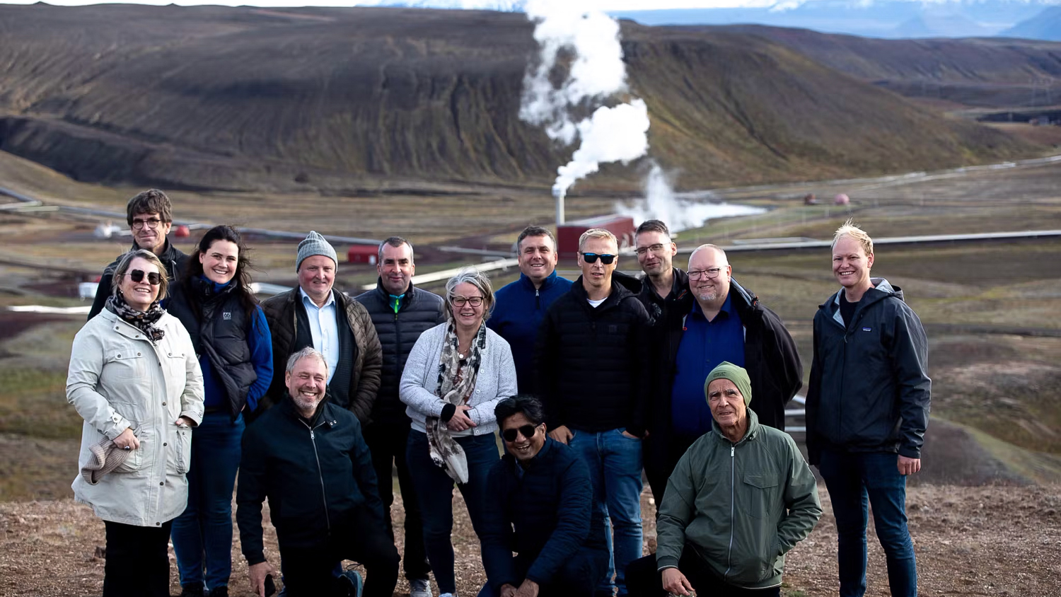 A group of sixteen people posing and smiling outdoors in a rugged, open landscape with geothermal steam rising and hills in the background. Most are wearing jackets, suggesting cool weather.