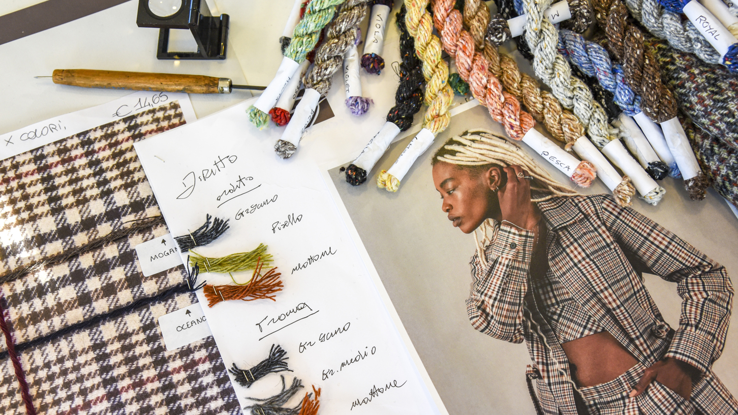 A designer’s workspace with color threads, fabric swatches, handwritten notes, and a fashion magazine featuring a model in a plaid suit with braided blond hair.