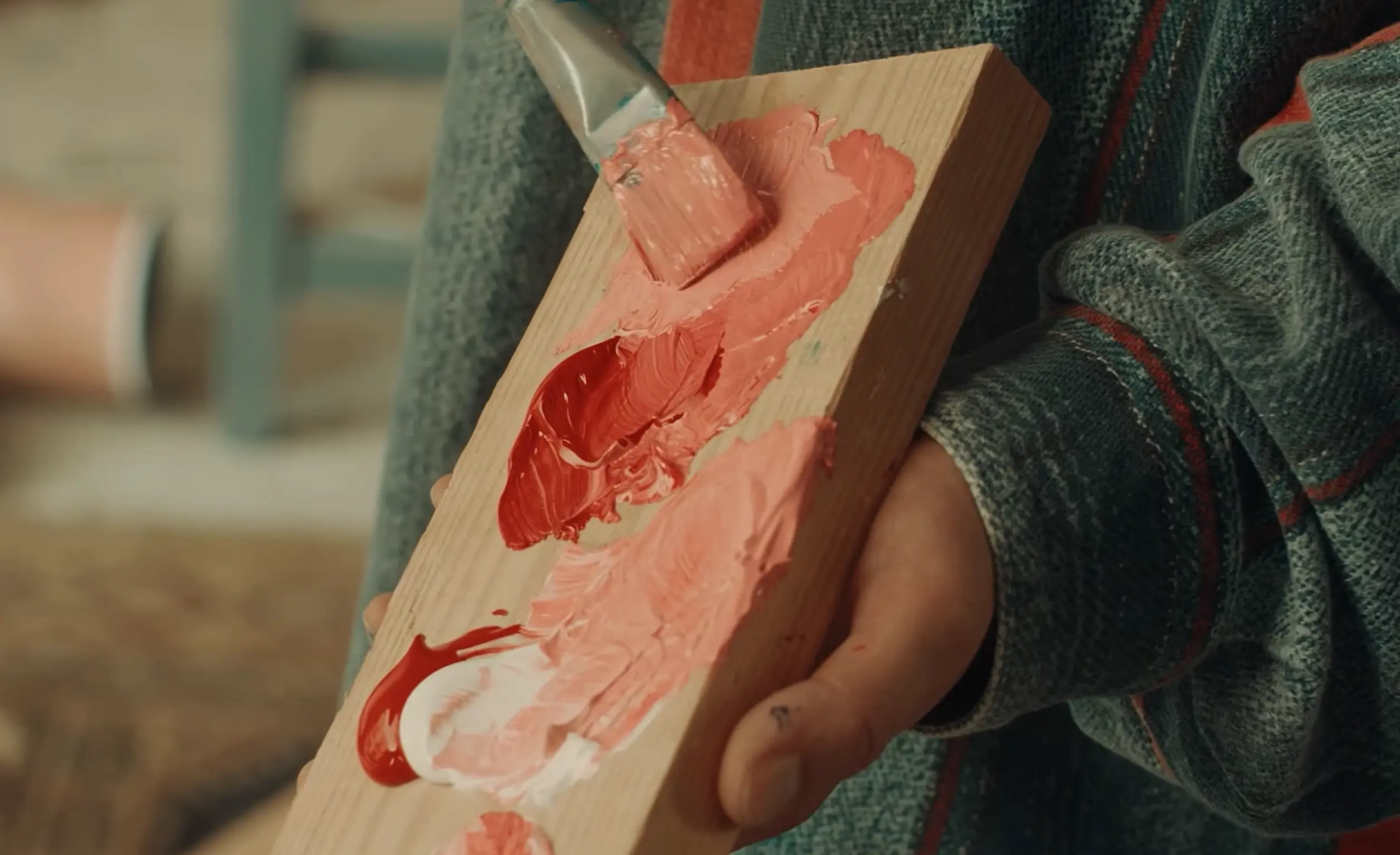 A person holds a wooden palette with various shades of red and pink paint, using a brush to mix the colors. The person is wearing a gray sweater with green stripes.