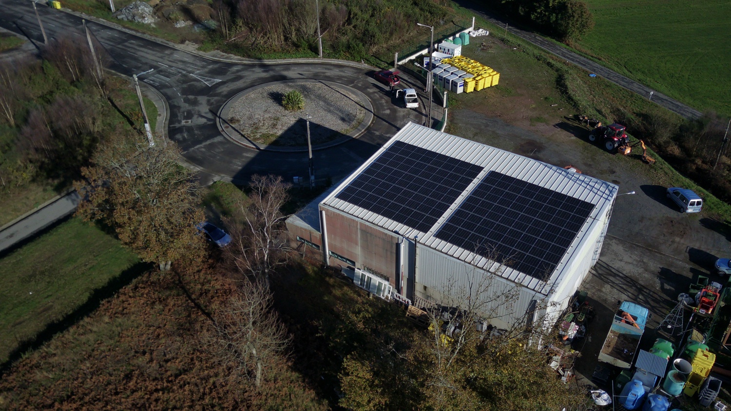Aerial view of a white industrial building with solar panels on the roof, surrounded by various equipment, trucks, and a roundabout, next to a grassy field and trees.