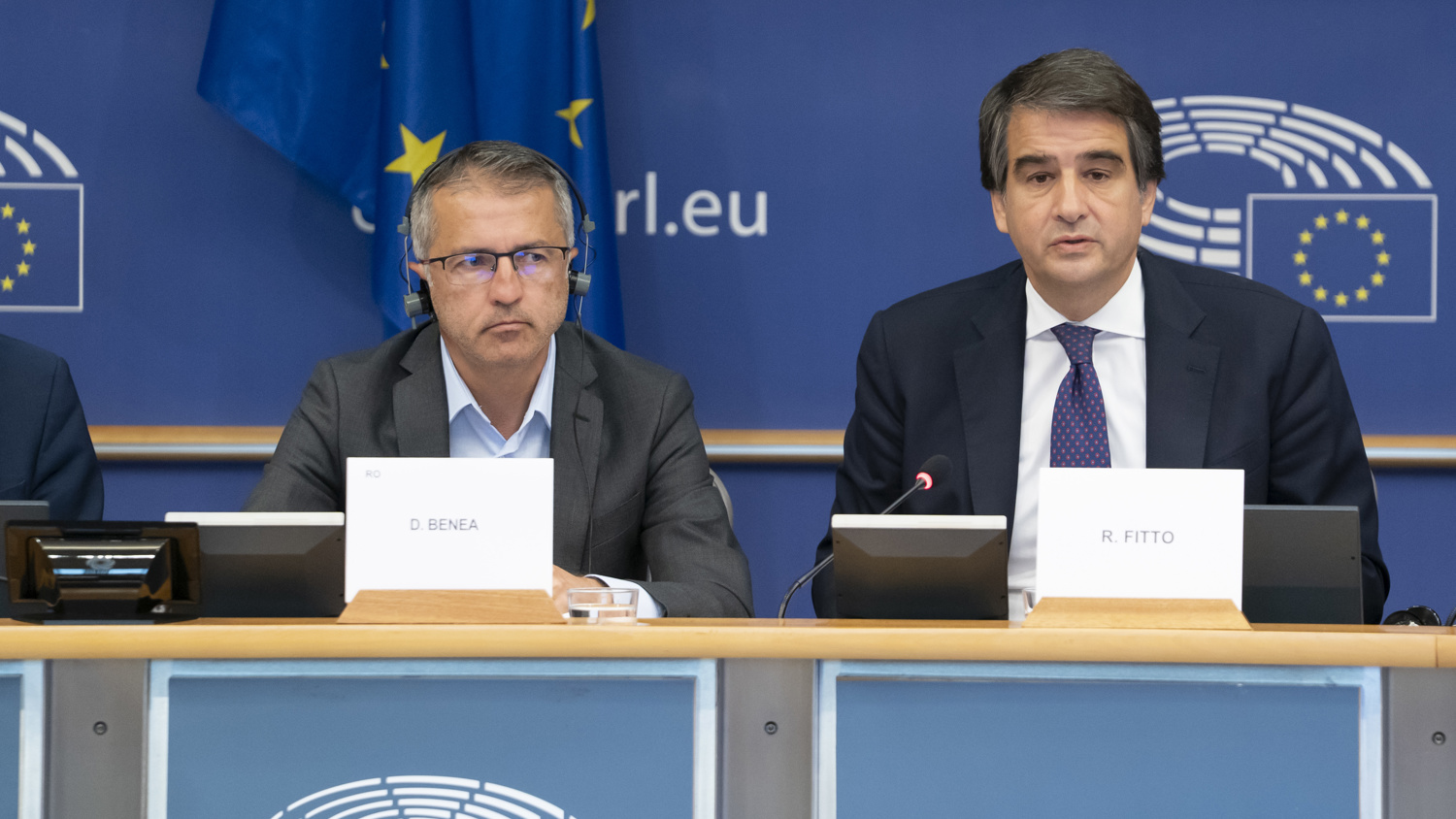 Two men in suits sit at a conference table with microphones, nameplates, and European Parliament logos in the background. Blue banners with the EU flag and the text "europarl.eu" are visible behind them.