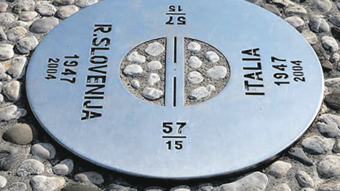 A round metal plaque set among cobblestones marks the border between Slovenia and Italy, with the years 1947 and 2004 and the words "R. Slovenija" on one side and "Italia" on the other.