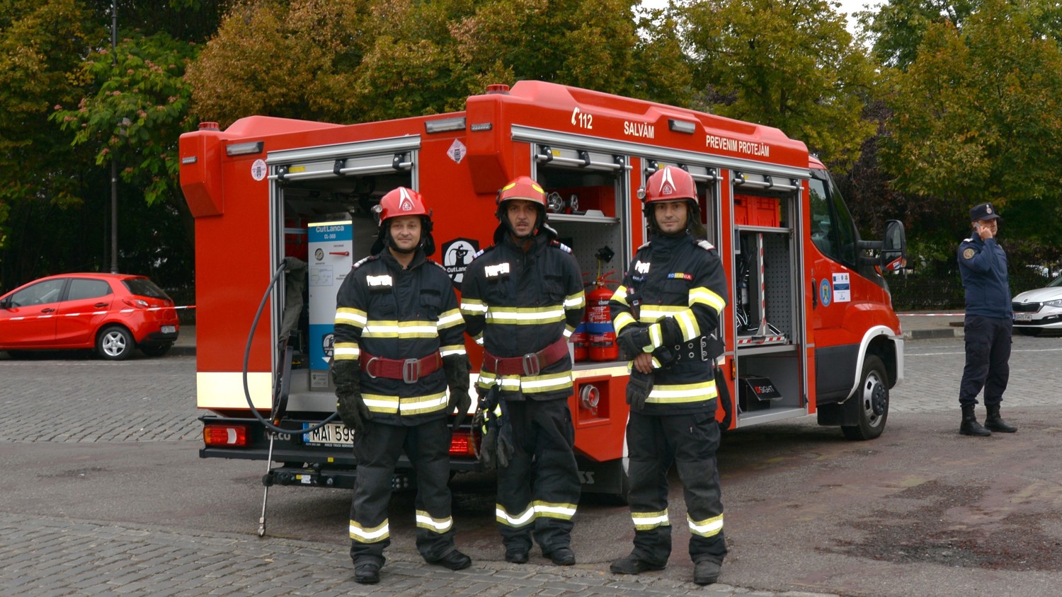 Three firefighters in uniform and helmets stand in front of a red fire truck with open compartments, while another person in uniform stands to the side. Trees and parked cars are visible in the background.