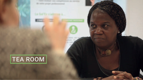 Two women sit across from each other at a table having a conversation. The words "TEA ROOM" appear on the image. The focus is on the woman with braided hair and a dark top, with the other woman blurred in the foreground.