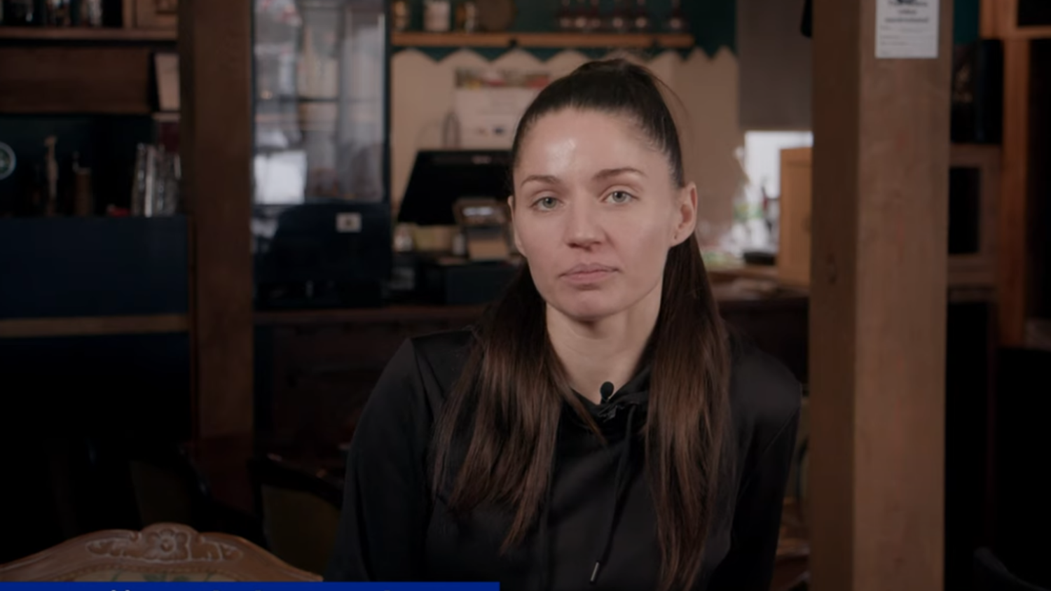 A woman with long dark hair tied back is sitting in a dimly lit room with wooden walls. She is wearing a black top and looking at the camera with a neutral expression. Various items are visible in the background, including a cash register.