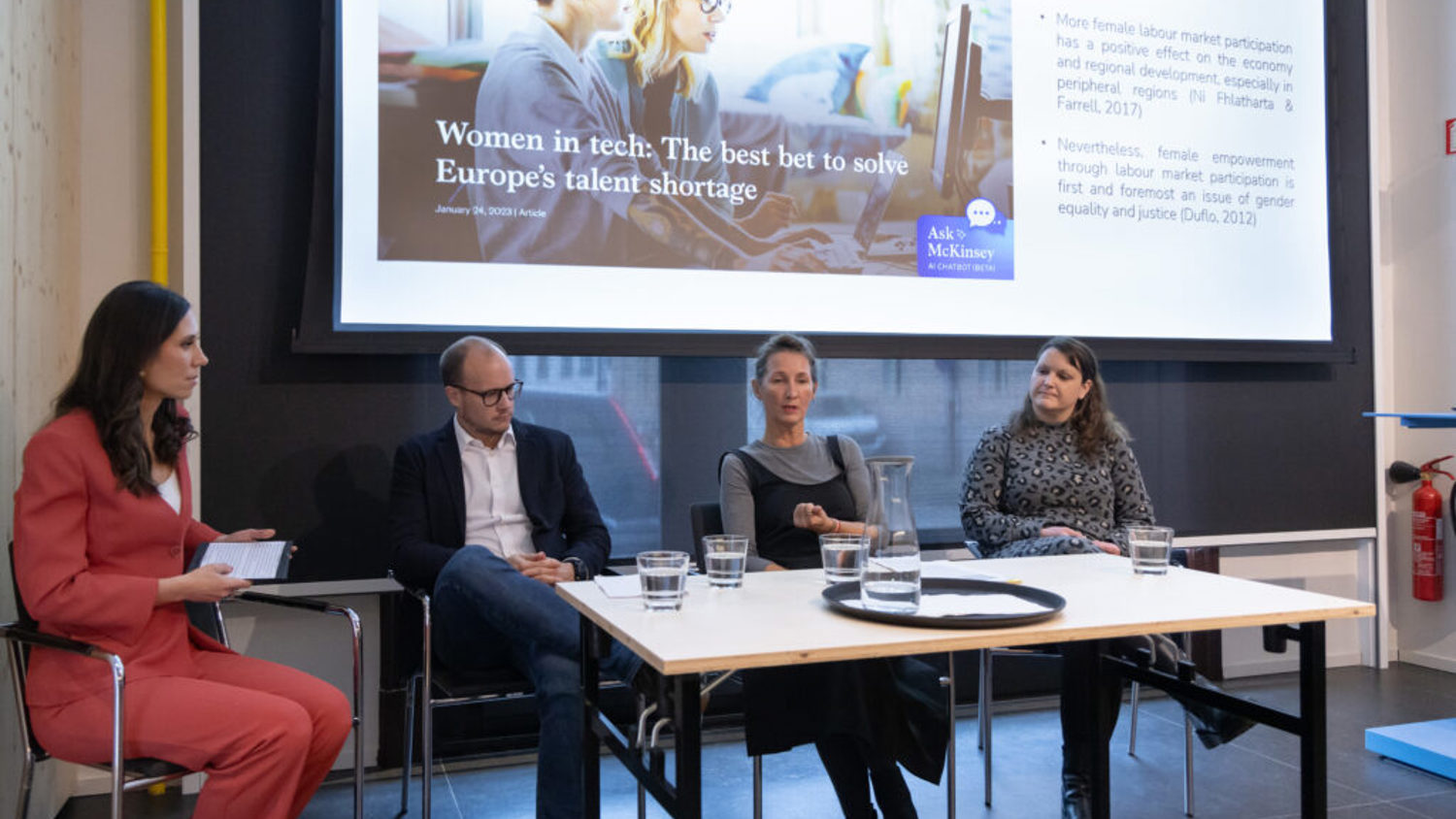 Four people sit on a panel in front of an audience, discussing women in tech. A screen behind them displays the topic: “Women in tech: The best bet to solve Europe’s talent shortage” with bullet points and an image of two women at a computer.