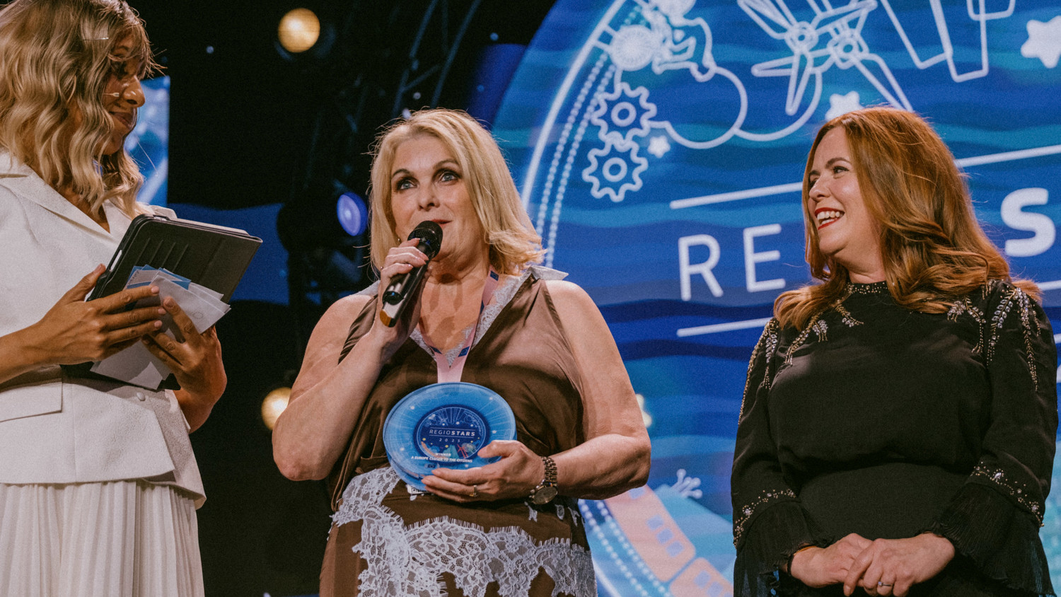Three women stand on stage at an award event. One holds a microphone and a blue glass award, another holds a notepad, and the third smiles. A large, colorful event logo is displayed in the background.