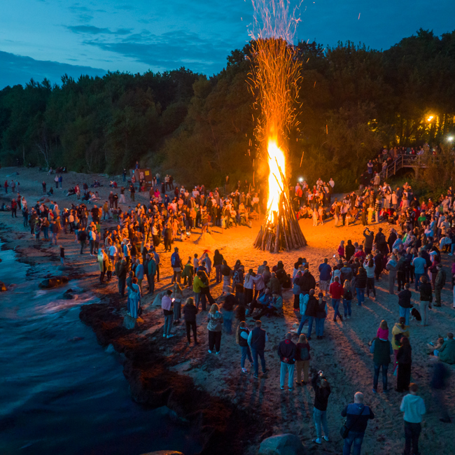 A large crowd gathers on a beach at dusk around a tall, blazing bonfire. The sea is on the left, and trees border the beach on the right. Sparks rise from the fire, and groups of people are scattered along the sand.